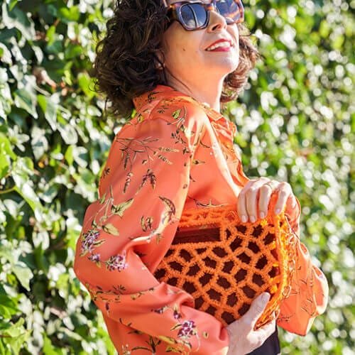 Mujer vestida con una chaqueta roja vibrante, bufanda colorida y pantalones oscuros, posando en una calle de Madrid. Lleva un bolso naranja de diseño 3D y una cadena dorada, combinando artesanía y moda contemporánea. El bolso destaca como un accesorio artesanal único que complementa su elegante look.